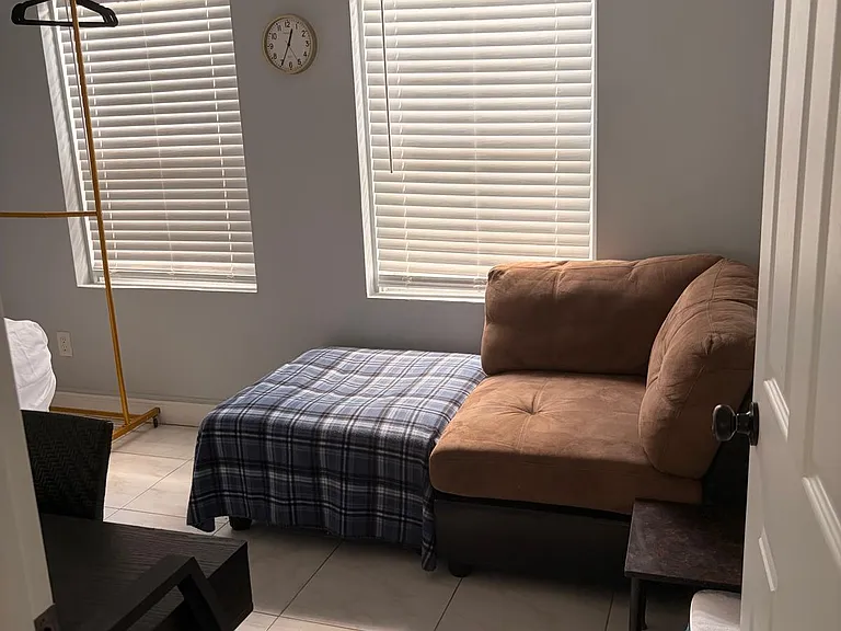 Compact bedroom with tile floors, two windows with blinds, and space for a bed plus seating and a side table.
