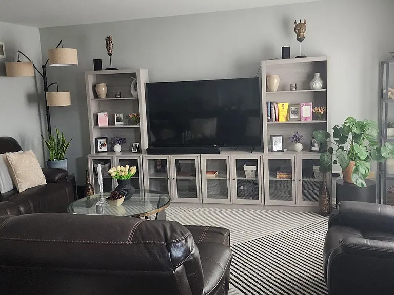Spacious living room with leather seating, large TV setup, display shelving, and natural light from nearby windows.