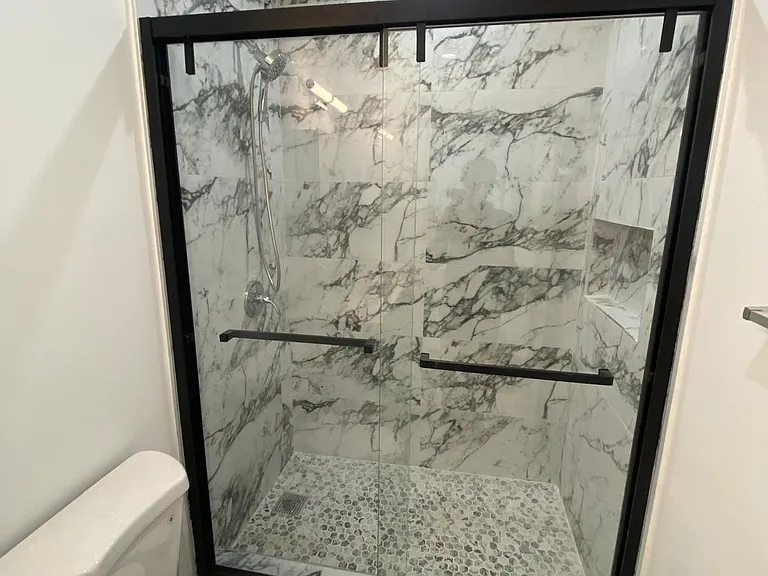 Walk-in shower with glass sliding doors, black trim, marble-look wall tile, built-in niche, and pebble-style shower floor.