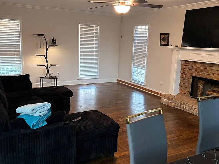 Open living area with hardwood floors, ceiling fan, and a stone fireplace, plus multiple windows for natural light.