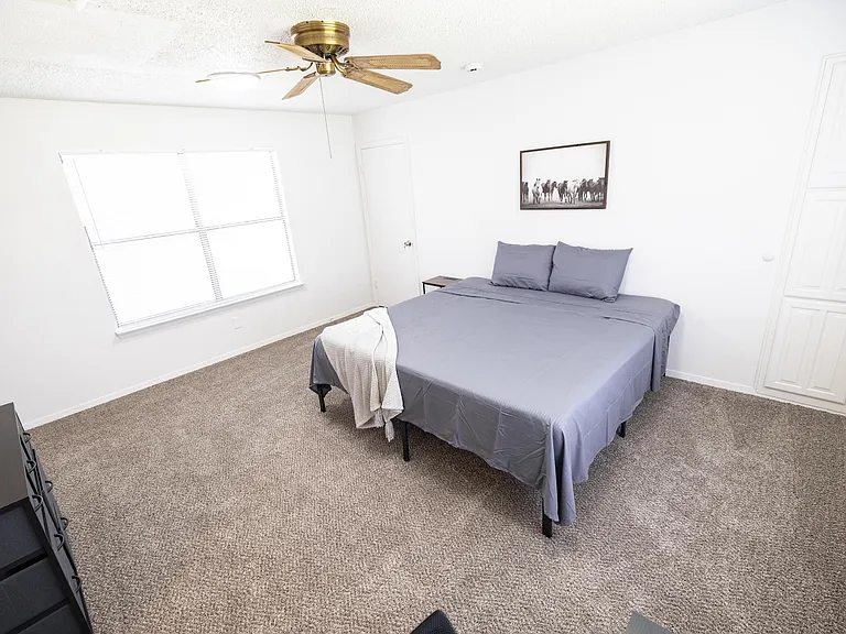 Carpeted bedroom with a queen bed, ceiling fan, large window with blinds, and closet storage for everyday convenience.