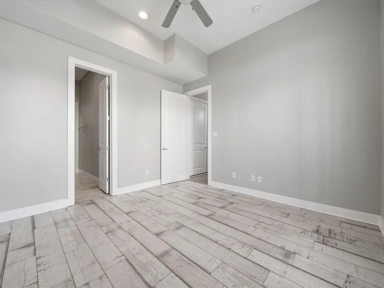 This downstairs bedroom offers a bright, open feel with light gray walls, wide wood-look flooring, and a ceiling fan. High ceilings with recessed lighting enhance the spacious layout, while dual access to the ensuite bathroom and walk-in closet