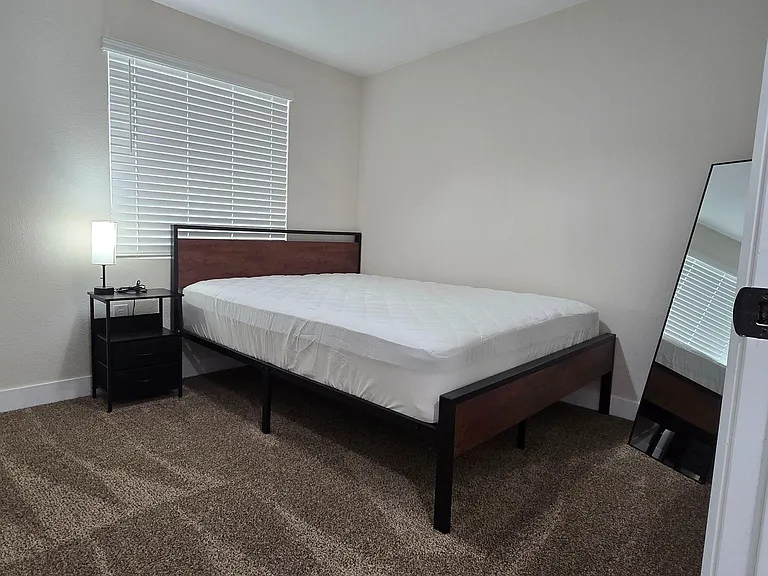 Furnished bedroom with a full-size bed, nightstand and lamp, plus a large window with blinds and carpeted flooring.