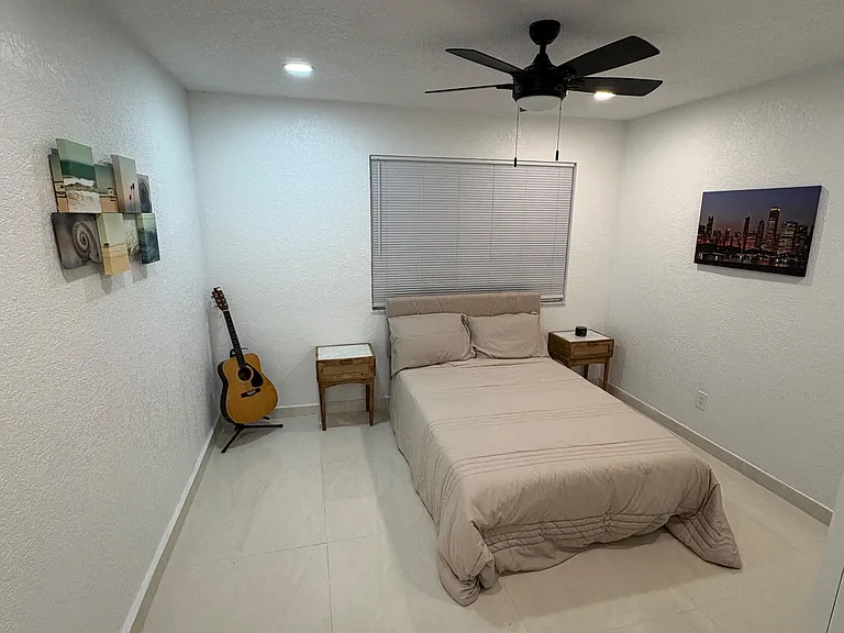 Private bedroom with tile flooring, ceiling fan, and a large window with blinds, plus space for bedside storage.