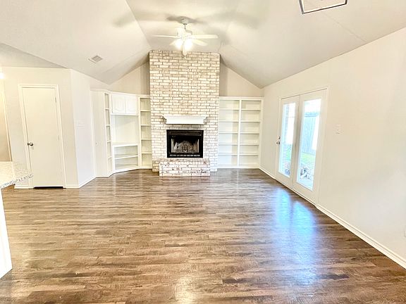Open living and dining room with vaulted ceiling, wood burning fireplace, and wood built-in shelving on either side of fireplace. Leads out to backyard through french door.
