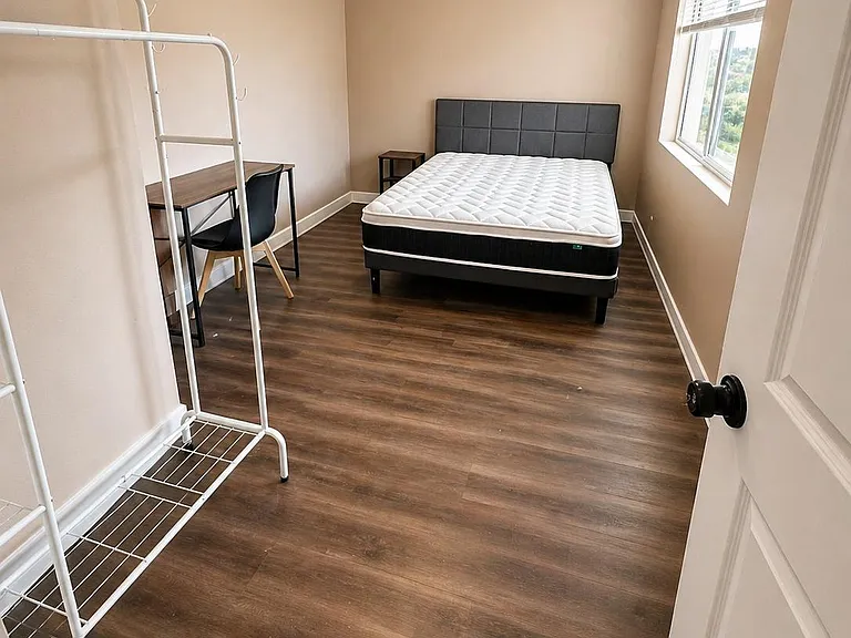 Furnished bedroom with wood-look flooring, a desk workspace, and a large window with blinds for natural light.