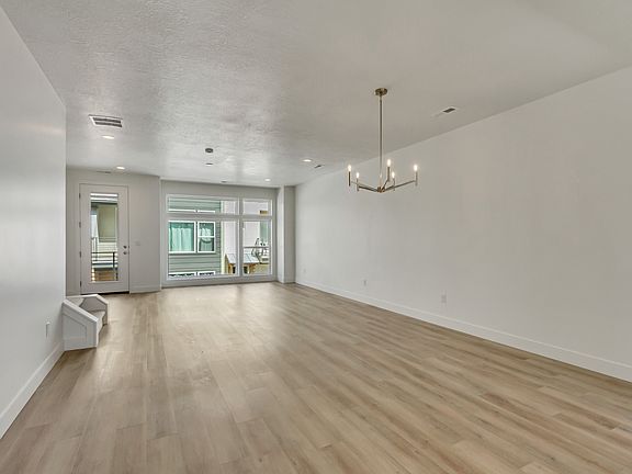 A spacious, empty room with light wood flooring, white walls, recessed lighting, a modern chandelier