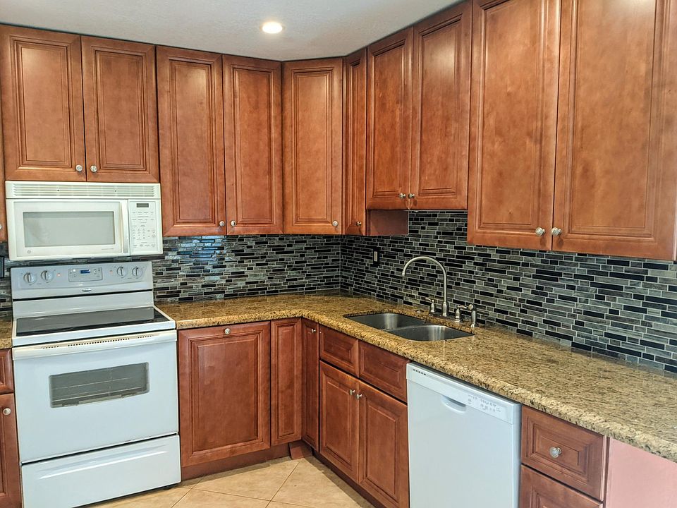 Corner kitchen with plenty of cabinet storage, double sink, and full appliance setup. Easy-to-clean tile backsplash and counters offer a functional layout for home cooks and meal preppers alike.