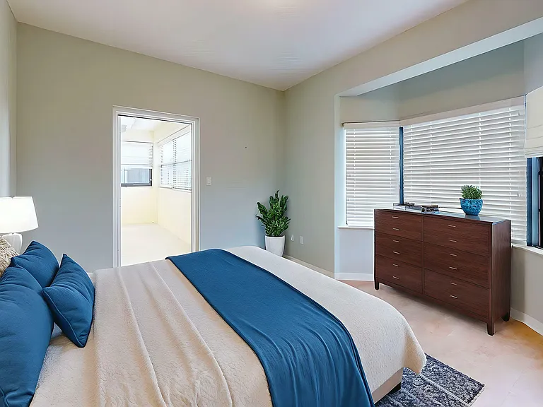 Inviting bedroom with plush bedding, elegant wooden dresser, large windows with blinds, and access to a bright sunroom.