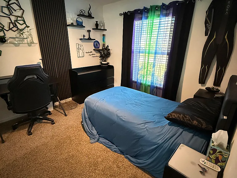 Carpeted bedroom with a ceiling fan, large window with blinds, and space for a bed, dresser, and desk setup.