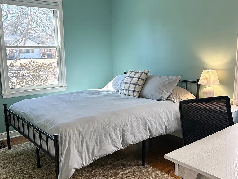 Main floor bedroom 2 features a queen size bed, desk and chair, nightstand, lamp, closet with a laundry basket and hangers.