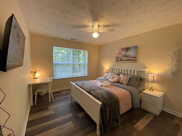 Staged bedroom with full bed, two nightstands, desk, and a large window with blinds plus ceiling fan lighting.