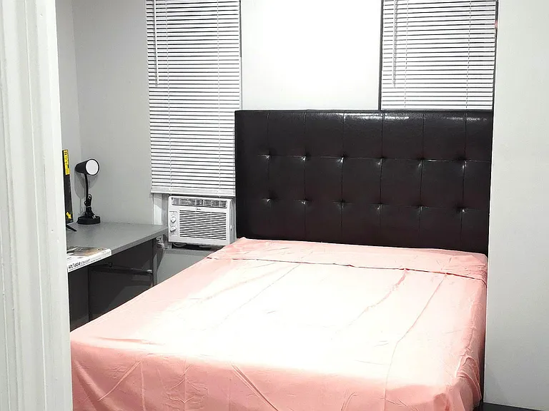 Furnished bedroom with a full bed, upholstered headboard, and a built-in desk area beside two windows with blinds.