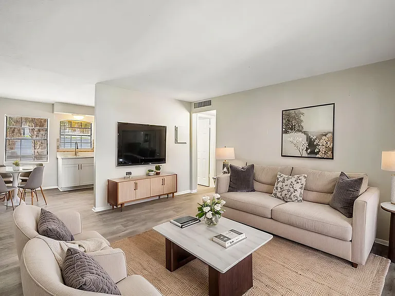 Bright, modern living room in an elegant apartment featuring cozy beige seating, a sleek entertainment center, and a charming dining area by a sunny window.