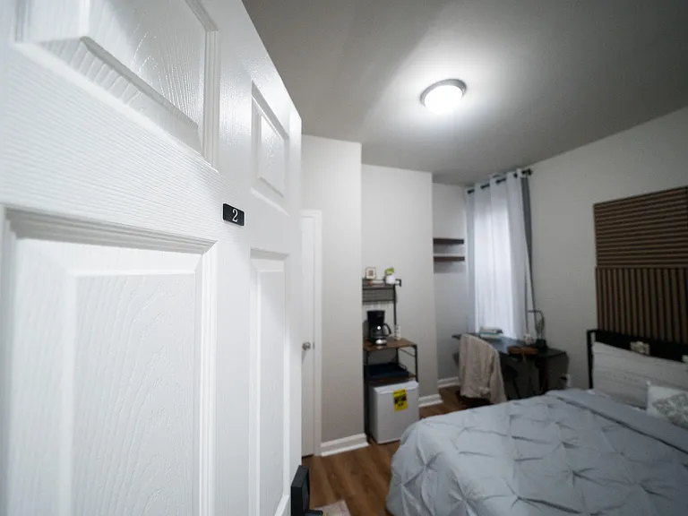 Private bedroom with a full bed, window with curtains, and a compact work area plus mini-fridge and coffee station.
