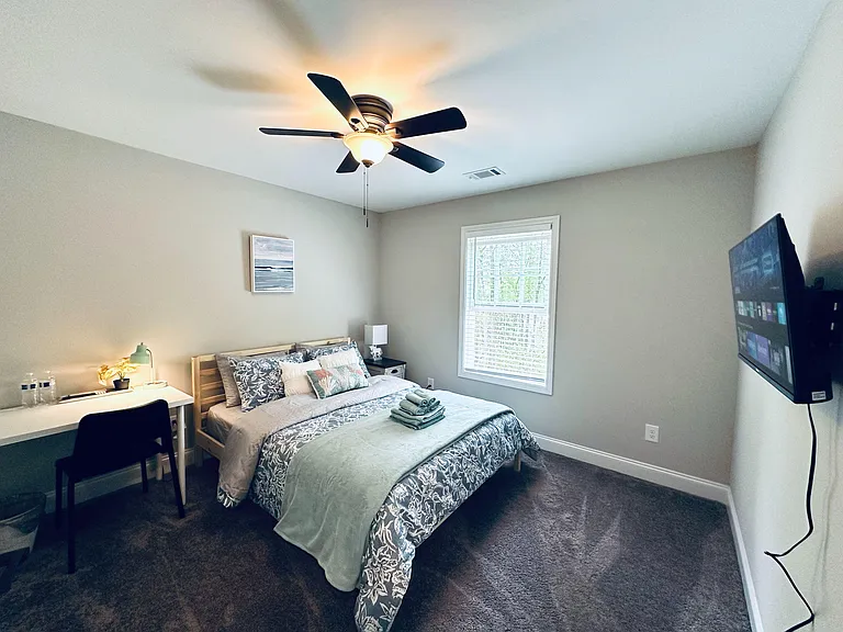 Furnished bedroom with a comfortable bed, bedside table, ceiling fan, and a work desk, plus a window with blinds.