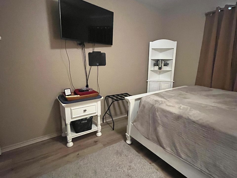 Furnished bedroom featuring a full bed, nightstand, and a wall-mounted TV, with a ceiling fan and window treatments.