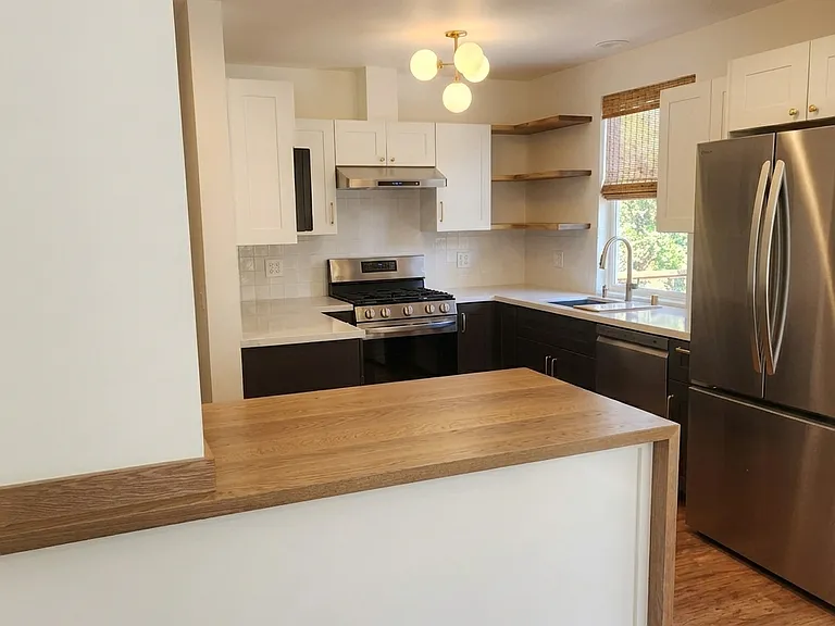 Kitchen view from the dining room. Custom hardwood waterfall island. All new appliances, cabinetry, solid wood open shelving, and custom woven-wood window shades. Kitchen sink view out to the back yard.