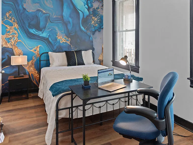 Furnished bedroom with a full bed, nightstand, and dedicated desk setup, plus large windows and hardwood-style flooring.