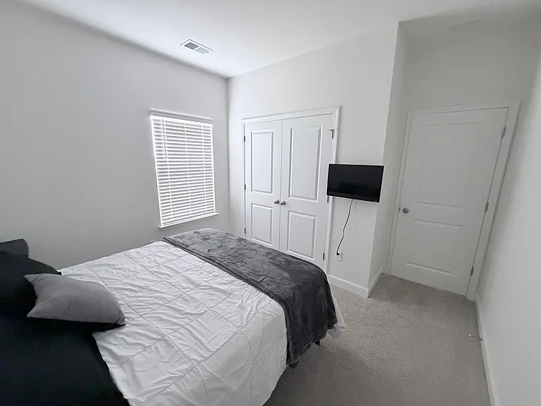 Furnished bedroom with a comfortable bed, wall-mounted TV, window with blinds, and a double-door closet for storage.