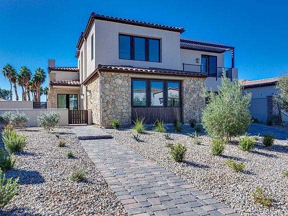 A modern, two-story Mediterranean-style house with a stone exterior, surrounded by a paved walkway a