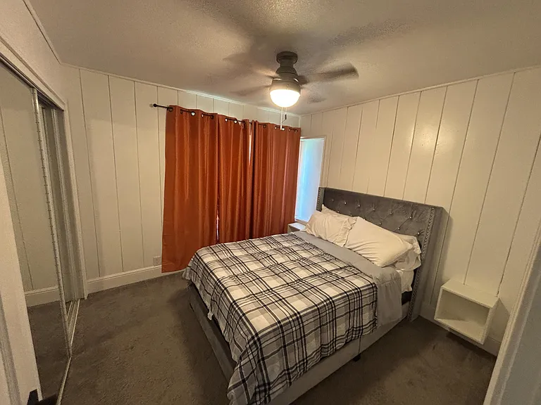 Private bedroom with a ceiling fan, paneled walls, and space for a full bed plus a nightstand; window with curtains for priva