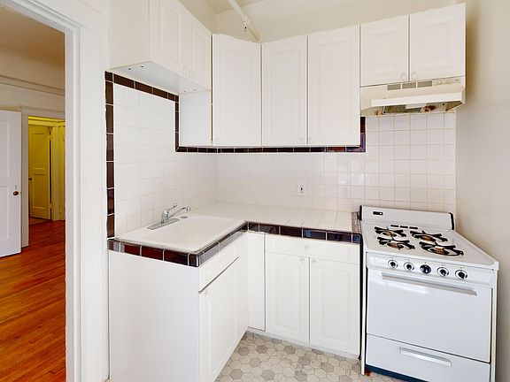 Compact kitchen with cabinets, trimmed backsplash, small sink, and a vintage gas stove. Hardwood flooring visible through door, cozy ambiance.