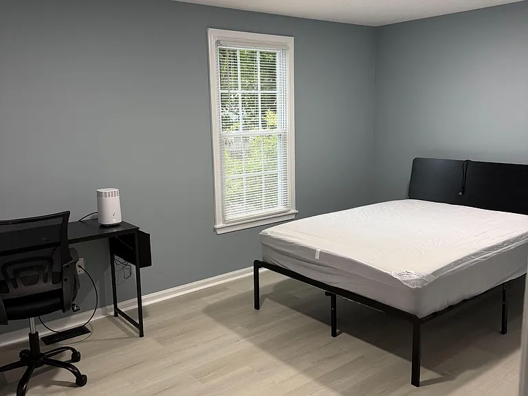Bedroom with neutral paint, large window with blinds, and durable flooring; includes space for a bed plus a small work area.