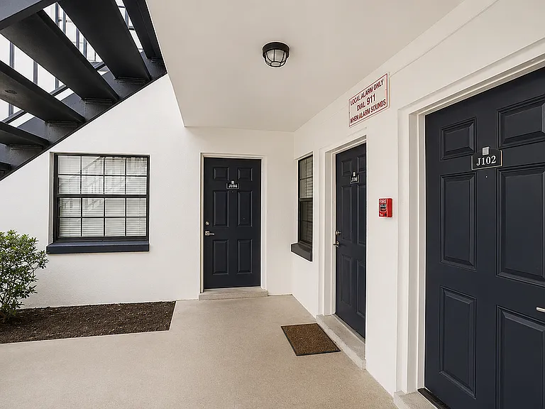 Modern apartment entrance with sleek black doors and a lush green shrub under an open staircase.