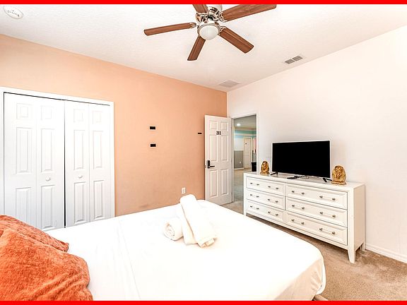 The room features a stylish upholstered bed, neutral carpeting, a ceiling fan, and ample natural light.