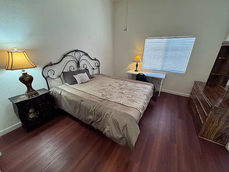 Furnished bedroom with a full bed, nightstand lighting, dedicated desk space, and wood-style flooring with window blinds.