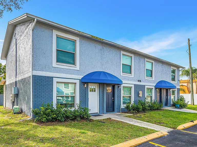 Charming double-story apartment with blue accents, featuring modern windows and lush landscaping under a clear sky.