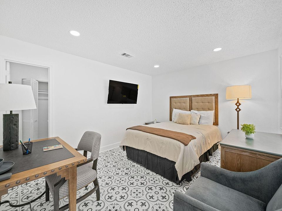 Furnished bedroom with queen bed, desk workspace, TV, and dresser, plus a closet and window with blinds.