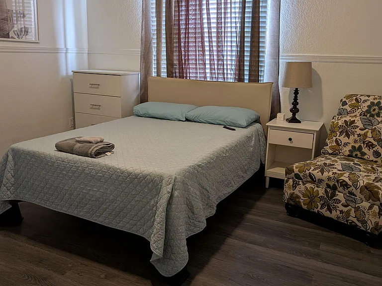 Furnished bedroom with a comfortable full-size bed, nightstand, dresser, and window with blinds for light control. TV