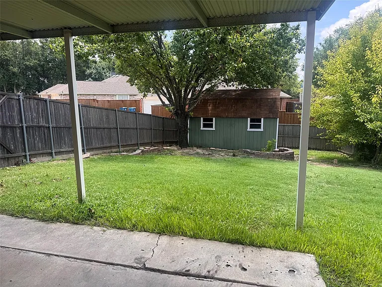 Covered patio opening onto a fenced backyard featuring a green shed with a brown roof, a mature tree, and a stone-lined fire pit