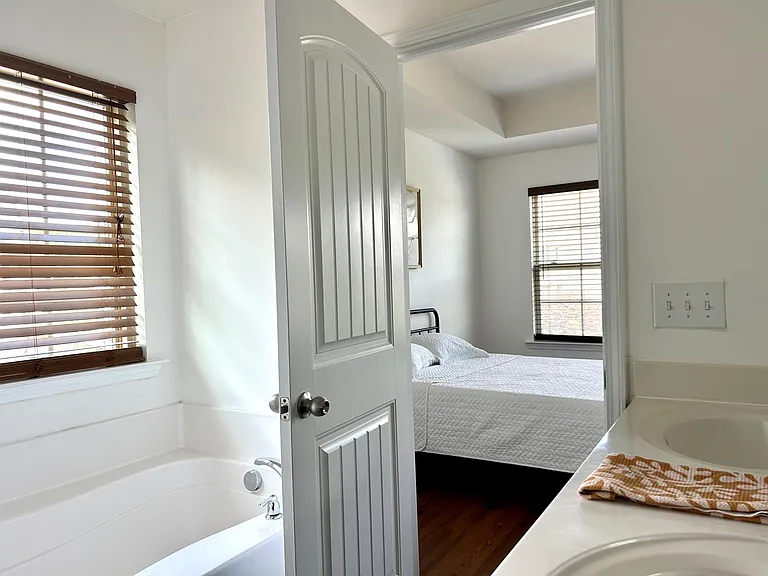 Full bathroom with tub/shower combo and long vanity counter, plus an adjacent bedroom visible through the doorway.