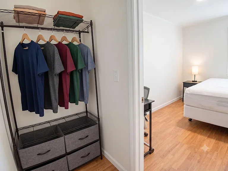 Bedroom includes a sleeping area with bedside lighting plus mini-fridge, open closet, and fabric drawers.
