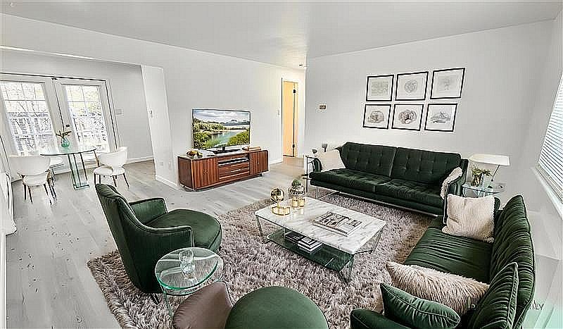 Elegant living room with plush green velvet sofas, a chic coffee table, and wall art; adjacent dining area with large windows letting in abundant natural light.