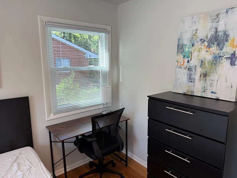 Bedroom area with a dedicated desk workspace, large window with blinds, and a dresser for convenient clothing storage.