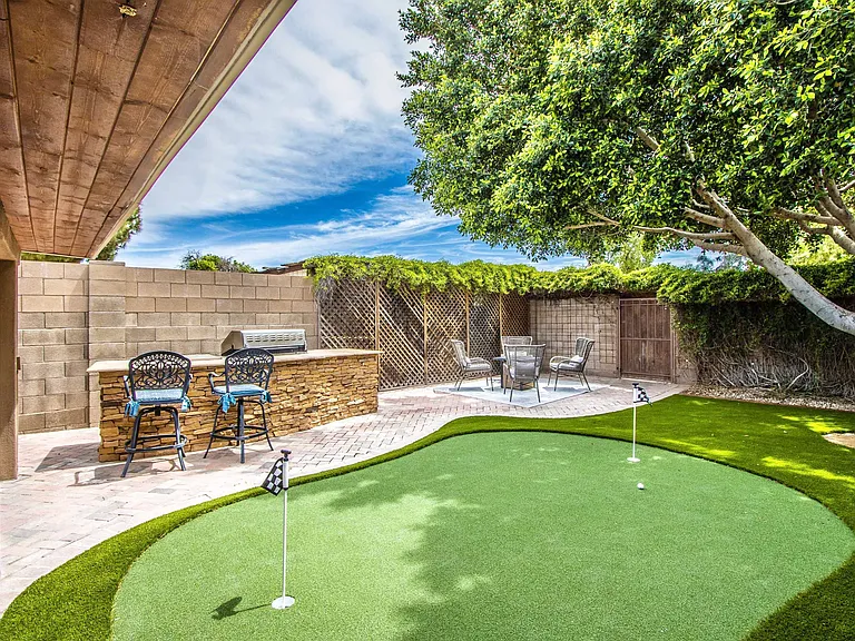 Full backyard view at Casa Del Sol, Tempe rental home. Features a custom putting green, stone BBQ island with bar seating, brick pavers, shade trees, and a patio fire pit set. The ultimate outdoor entertainment space for a top Tempe, AZ stay.