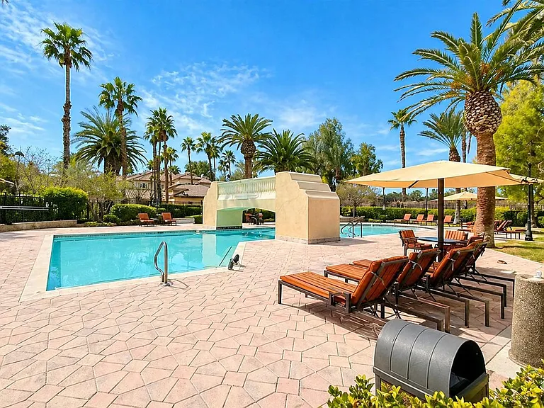 Resort-style outdoor pool area with ample lounge seating, shade umbrellas, and a spacious paver deck framed by palms.