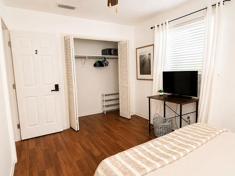 Bedroom with wood-look flooring, open closet storage, and a TV setup by the window for relaxing or working.