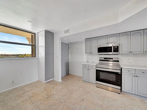 View of open kitchen from refrigerator side. New stainless steel stove and microwave. Storage pantry with deep shelves next to front window. All newly painted.
