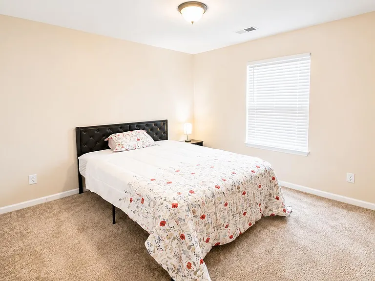 Spacious bedroom with carpeted floors, a large window with blinds, and room for a full or queen bed plus nightstands.