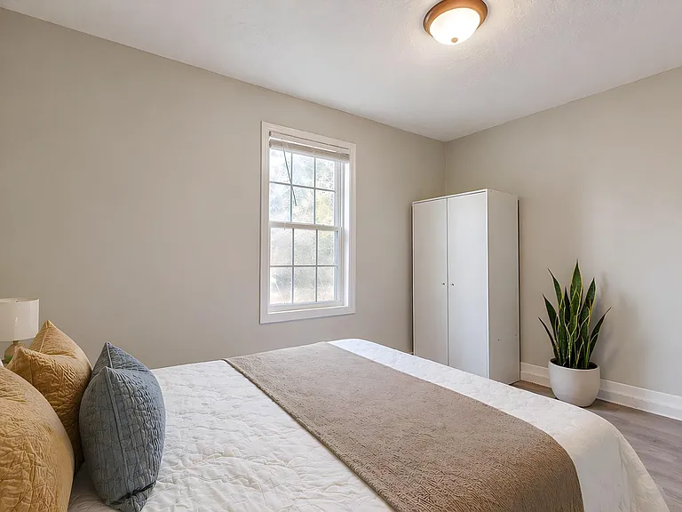 Private bedroom with a large window, neutral walls, and space for a bed plus wardrobe storage and a nightstand.
