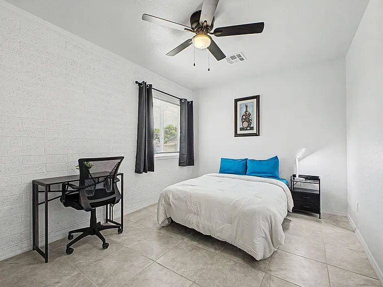 Private bedroom with a full bed, nightstand, and dedicated desk workspace, plus a window with curtains and ceiling fan.