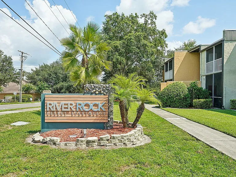 Inviting entrance to River Rock Luxury Apartments surrounded by lush greenery and palm trees on a sunny day.