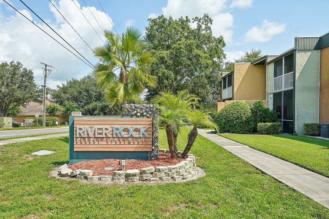 Inviting entrance to River Rock Luxury Apartments surrounded by lush greenery and palm trees on a sunny day.