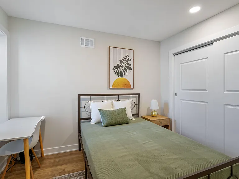 Furnished bedroom with a full bed, nightstand, and dedicated desk area, plus a sliding-door closet for storage.