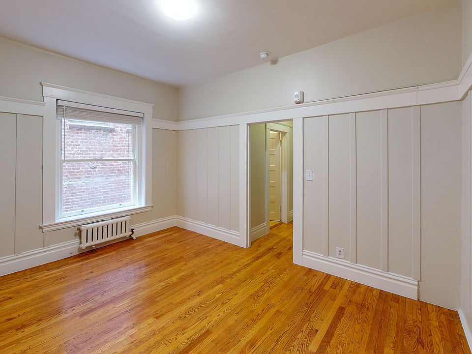 Bright, spacious room with hardwood floors, paneled walls, and a large window overlooking exposed brick, leading to a hallway.